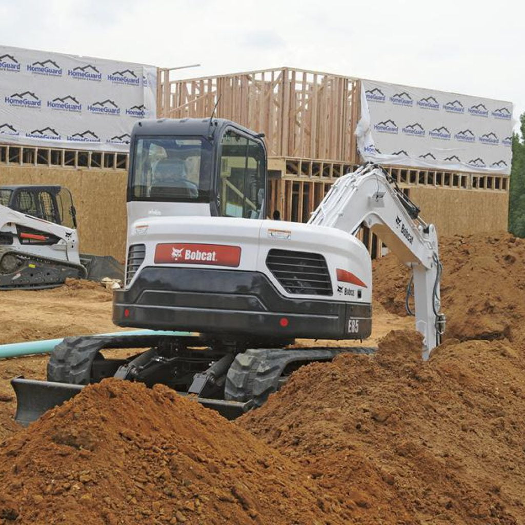 Mini excavadora Bobcat E35 en obra de construcción, moviendo tierra en un sitio de obras con maquinaria pesada y estructura en construcción en segundo plano.