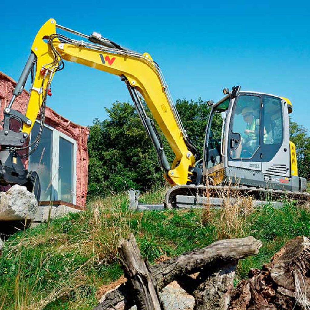Excavadora compacta en obra de construcción junto a estructura de ladrillos amarillos y vegetación.