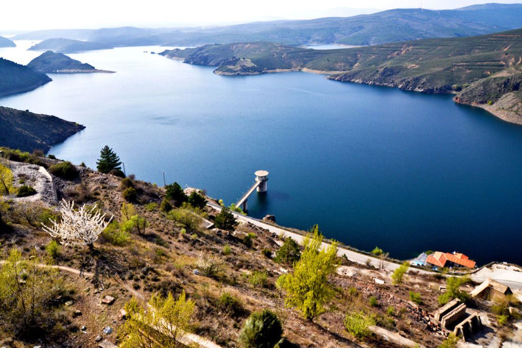 Imagen de un embalse de gran tamaño rodeado de montañas y vegetación, con una torre de control de agua a orillas del embalse, en un entorno natural y rural.