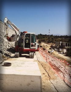Maquinaria de excavadora compacta trabajando en proyecto de construcción de carreteras en zona rural, con cielo azul y terreno rocoso, perfecta para obras de infraestructura y paisajismo.