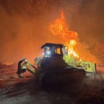 Maquinaria pesada en combate contra incendio forestal con una bulldozer en acción en un escenario nocturno, con llamas y humo intenso.